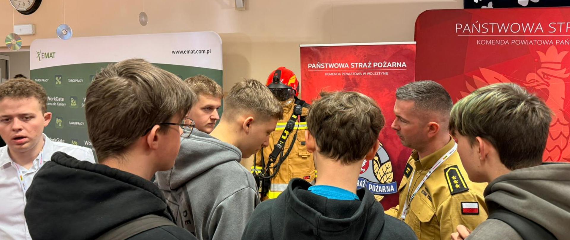 Grupa uczniów zgromadzona wokół funkcjonariusza Państwowej Straży Pożarnej, który opowiada o służbie i prezentowanym sprzęcie. W tle znajdują się roll-upy promocyjne PSP oraz manekin ubrany w pełne ubranie specjalne strażaka.