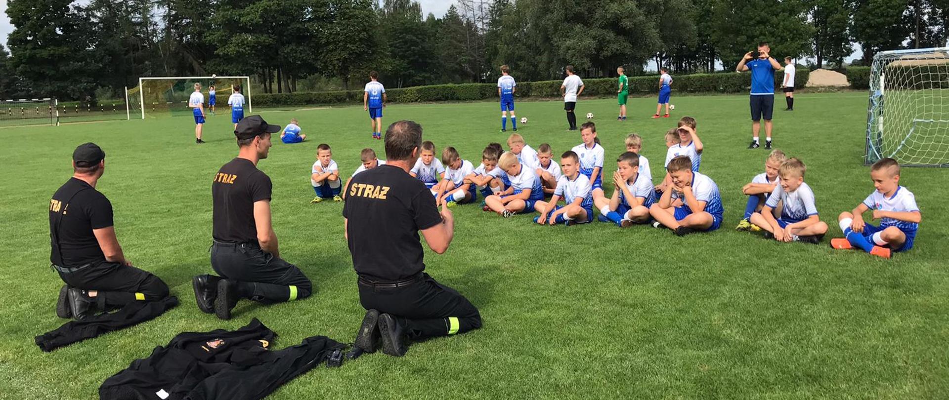 Zdjęcie przedstawia młodzieżową drożynę piłkarską, która siedzi na murawie boiska i słucha wykładu strażaków na temat bezpieczeństwa, jedne z chłopców nie słucha strażaków i ma odwróconą głowę w przeciwnym kierunku. W tle widzimy bramki oraz grających chłopców.