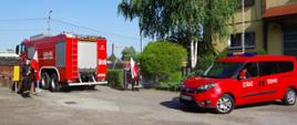 Wyjazd z placu JRG pojazdów PSP ze strażakami udającymi się do Grecji. Po lewej stronie pojazd typu GCBARt, za nim pojazd typu SLKw. W bramie wyjazdowej stoją dwaj strażacy z biało-czerwonymi flagami.