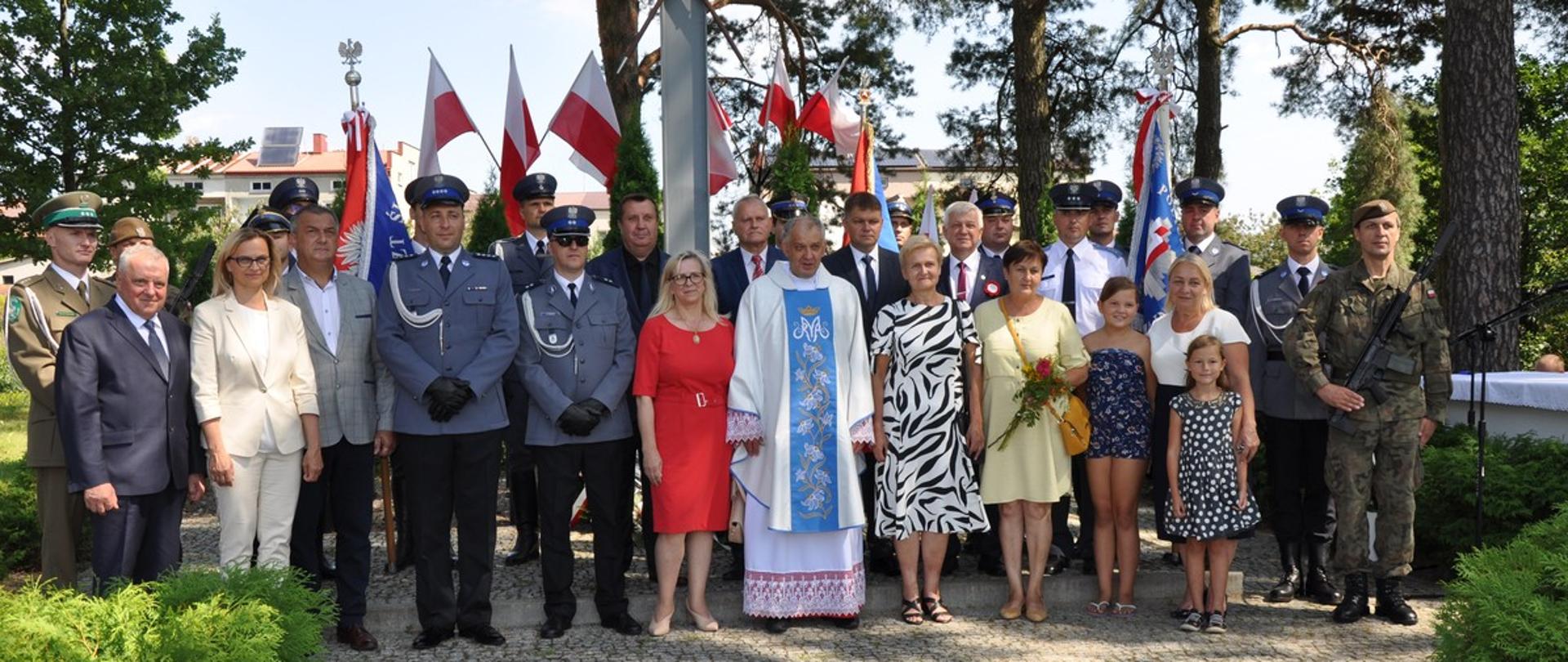 Na tle biało-czerwonych flag stoją w dwóch rzędach przedstawiciele samorządów, służby mundurowe ( Policja, Straż Pożarna, Straż Graniczna Służba Więzienna oraz Wojsko Polskie). Po środku stoi kapłan w ubraniu liturgicznym. 