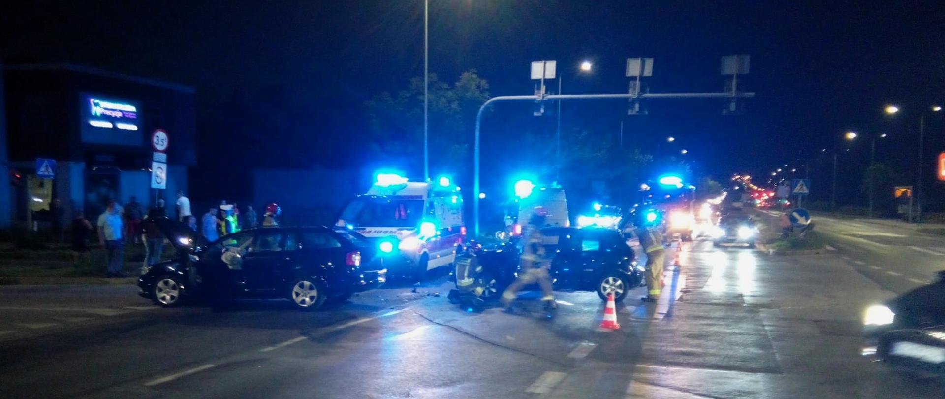 Zdjęcie przedstawia miejsce wypadku: w centralnym punkcie widzimy 2 samochody stojące na 2 pasach jezdni, wśród nich przechodzą ratownicy, a w głębi widać 2 ambulanse oraz samochody pożarnicze.