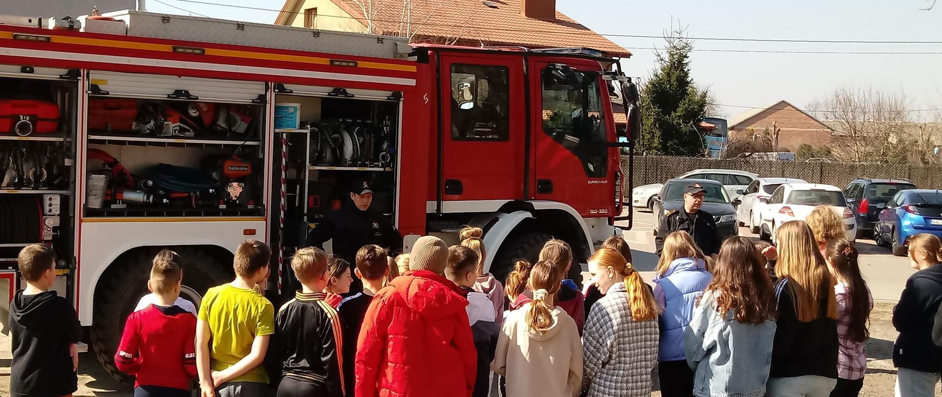Na zdjęciu widoczny jest czerwony samochód pożarniczy z otwartymi skrytkami sprzętowymi. W środku znajduje się sprzęt ratowniczo-gaśniczy. Przy samochodzie zgrupowane są dzieci. Dzieci w różnym wieku słuchają dwóch strażaków opowiadających o sprzęcie oraz zawodzie strażaka. W tle widoczne jest błękitne niebo oraz zabudowania.