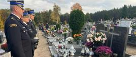 Zdjęcie przedstawia strażaków na cmentarzu, składających znicze przy grobie.
W tle nagrobki, drzewa.
