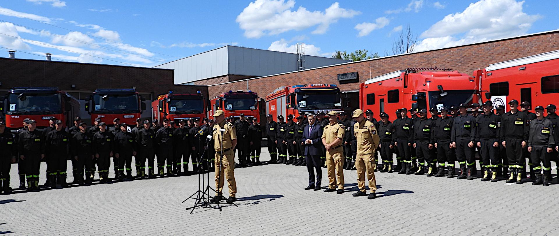 zastępca komendanta głównego Państwowej Straży Pożarnej nadbryg. Arkadiusz Przybyła przemawia do mikrofonów na środku placu, za nim stoją: wojewoda śląski Jarosław Wieczorek, śląski komendant wojewódzki nadbryg. Jacek Kleszczewski oraz dowódca grupy bryg. Grzegorz Borowiec. Wokół nich stoją strażacy w czarnych mundurach koszarowych. W tle samochody straży pożarnej i budynek jednostki.