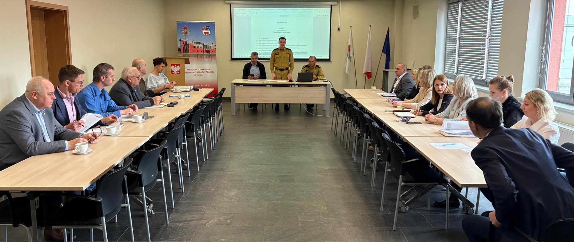 Uczestnicy spotkania siedzą przy stołach, słuchając przemawiającego Komendanta Powiatowego Państwowej Straży Pożarnej w Turku. 