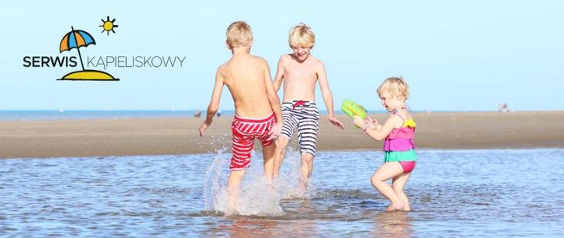 Dzieci bawią się na plaży w wodzie. W lewym górnym roku logo parasol wbity w piasek i słońce.