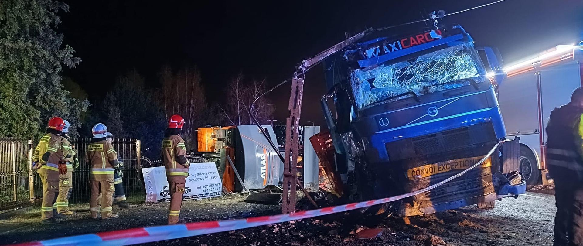 Samochód ciężarowy po wypadku, na którym leży złamany słup energetyczny. Obok stoją strażacy. W tle samochód strażacki.
