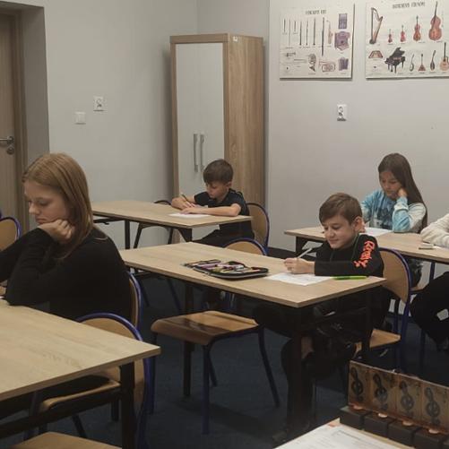 Zdjęcie przedstawia uczniów siedzących w ławkach i rozwiązujących test mający wyłonić laureatów I Szkolnego Konkursu ,, Mistrz Dyktanda", w tle ilustracje przedstawiające instrumenty. 