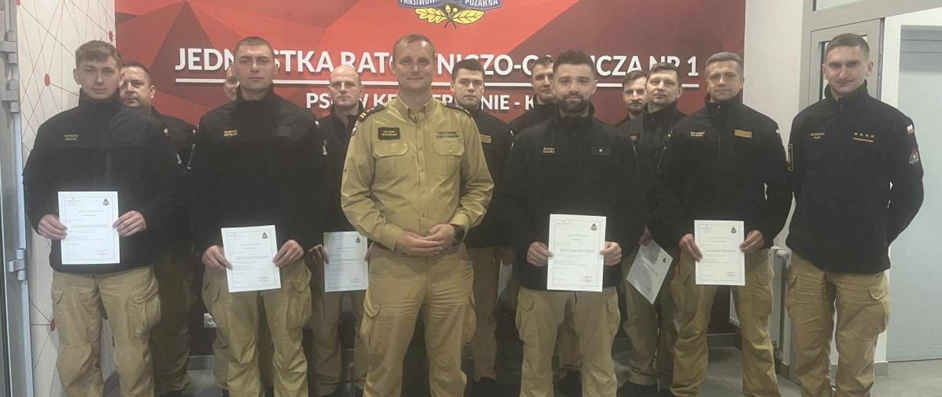 Grupa funkcjonariuszy PSP stoi w pomieszczeniu przed ścianą z czerwonym tłem i emblematem jednostki „JRG nr 1 Państwowa Straż Pożarna”. Kilku strażaków trzyma w dłoniach certyfikaty. Zdjęcie przedstawia uroczyste zakończenie szkolenia doskonalącego.