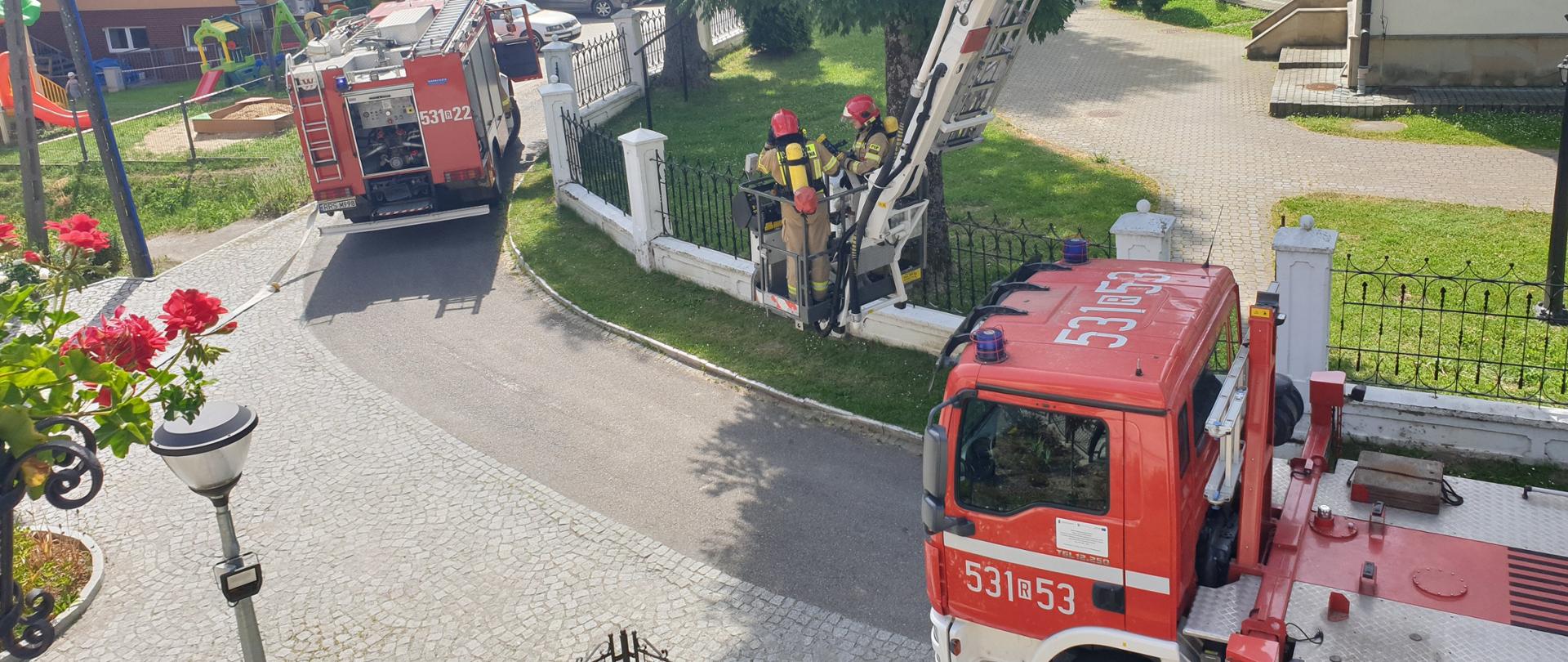 Zdjęcie wykonane z okna budynku. W tla dwa samochody pożarnicze. jeden to podnośnik hydrauliczny. W koszu podnośnika stoją dwa strazacy ubrani w ubrania bojowe.
