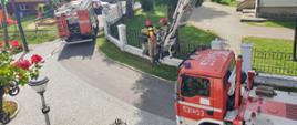 Zdjęcie wykonane z okna budynku. W tle dwa samochody pożarnicze. jeden to podnośnik hydrauliczny. W koszu podnośnika stoją dwa strażacy ubrani w ubrania bojowe.