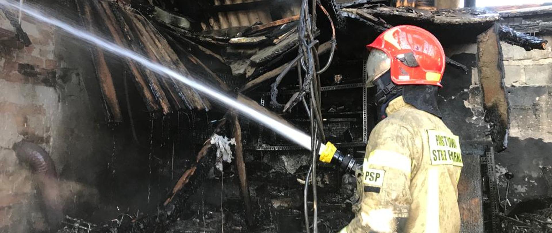 Strażak stojący po prawej stronie polewa zgliszcza budynku gospodarczego w lewą stronę.