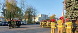 Trzy grupy strażaków w ubraniach bojowych stojących na placu przed budynkiem komendy. Jedna grupa trzyosobowa stoi przy maszcie. W tle drzewa i budynki.