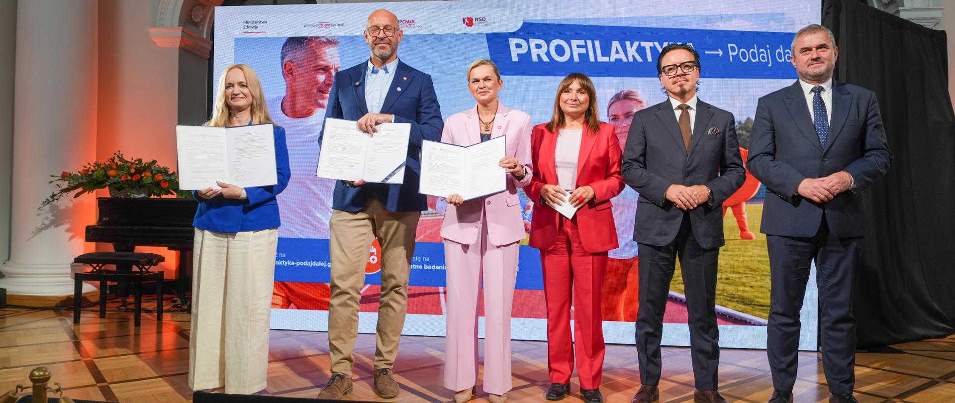 Sześć osób w eleganckich strojach stoi w rządku na scenie przed wielkim telebimem z hasłem 'PROFILAKTYKA Podaj dalej'. Trzy osoby po lewej stronie trzymają w rękach otwarte teczki z podpisanymi dokumentami.