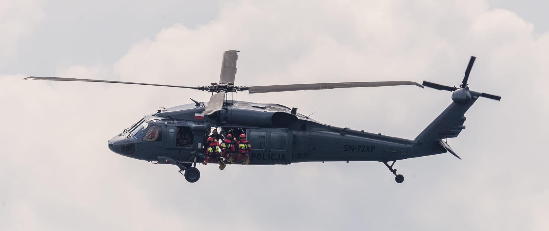 W powietrzu leci policyjny śmigłowiec, z którego właśnie wychylają się do zejścia na linie w dół 3 ratownicy wysokościowi