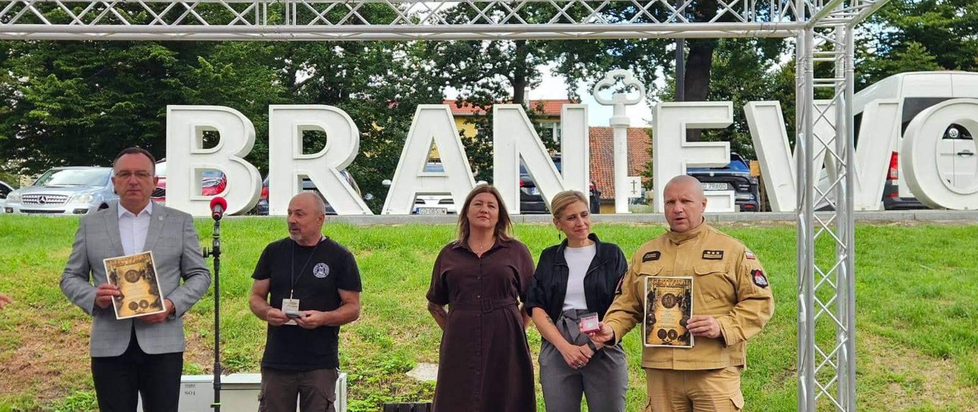 Na zdjęciu uwieczniono uroczyste wręczenie wyróżnień podczas wydarzenia w Braniewie. Na scenie stoją przedstawiciele organizatorów i zaproszeni goście, w tym strażak w umundurowaniu, którzy prezentują pamiątkowe dyplomy i odznaczenia. W tle widoczny jest charakterystyczny napis „BRANIEWO”, podkreślający miejsce wydarzenia.