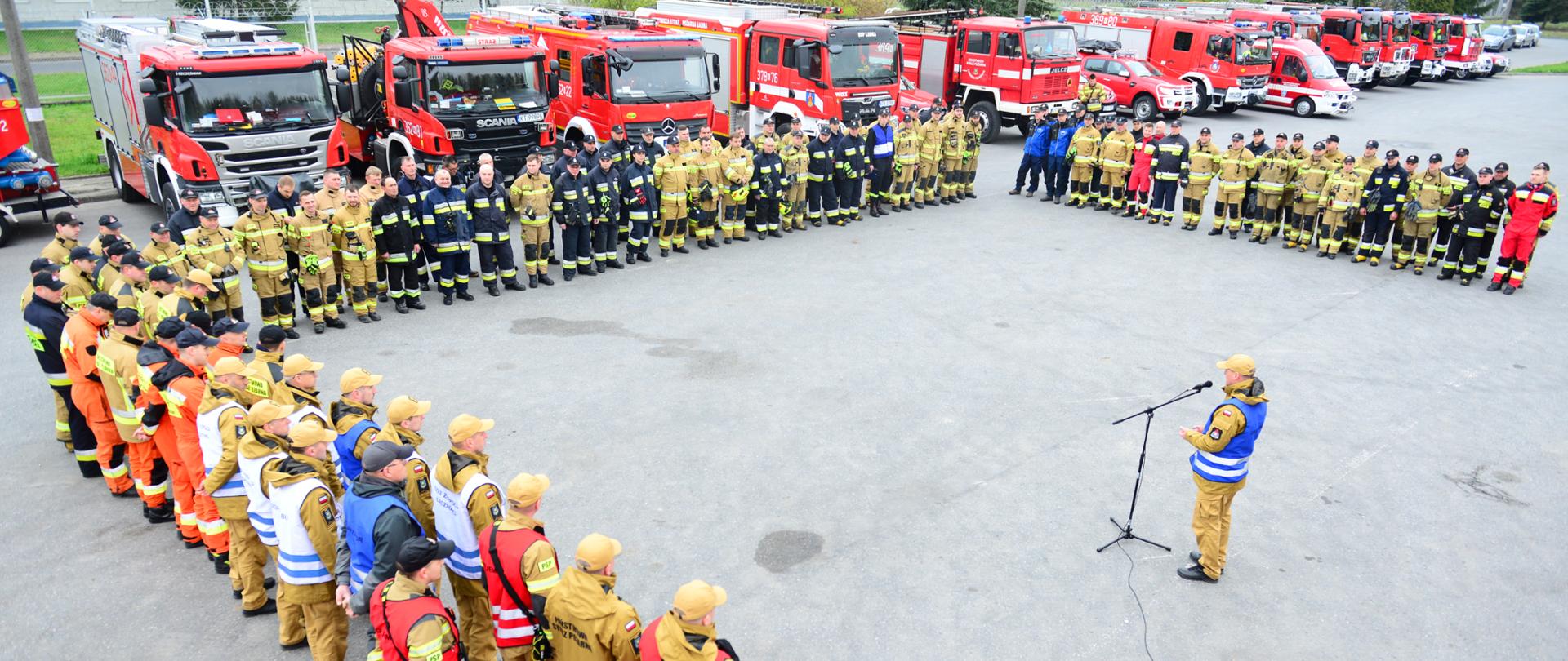 Na zdjęciu widać strażaków podczas odprawy przed ćwiczeniami. Strażacy stoją w układzie litery U. Ubrani są w ubrania specjalne na środku kierownik ćwiczeń i przemawia do ratowników 