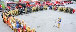 Na zdjęciu widać strażaków podczas odprawy przed ćwiczeniami. Strażacy stoją w układzie litery U. Ubrani są w ubrania specjalne na środku kierownik ćwiczeń i przemawia do ratowników 