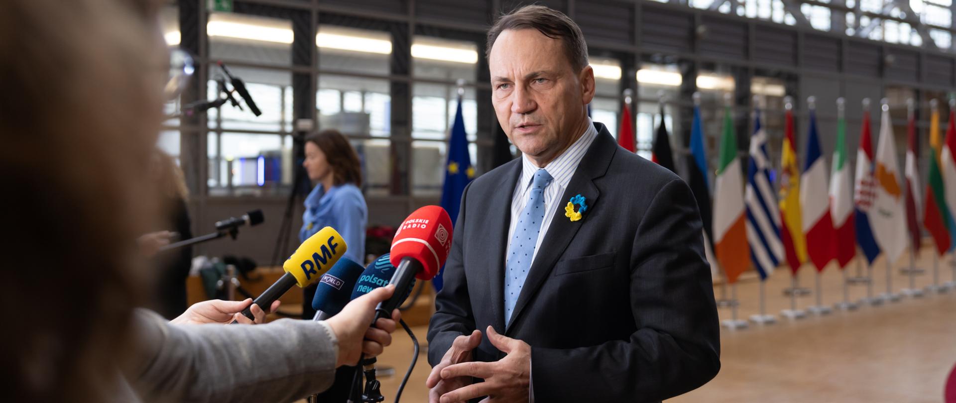 Mężczyzna w garniturze i krawacie udziela wypowiedzi mediom w przestrzeni konferencyjnej; przed nim widoczne są mikrofony i dyktafony dziennikarzy, w tle stoją flagi państw.