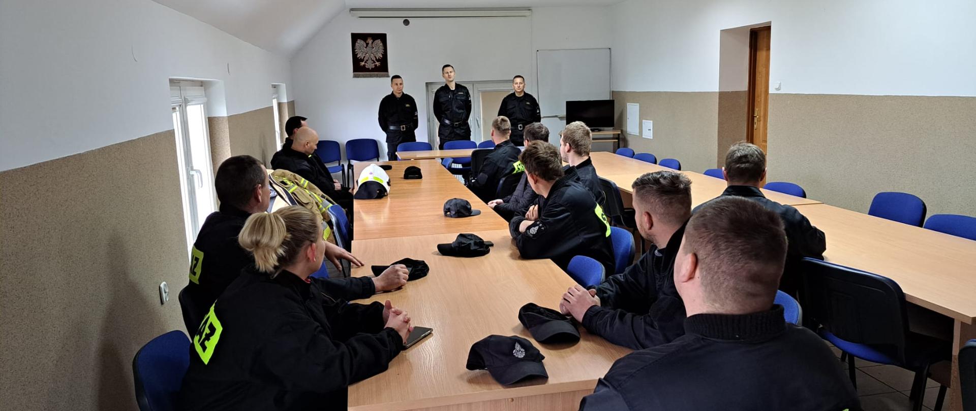 Zdjęcie przedstawia strażaków OSP ubranych w ubrania koszarowe koloru czarnego z napisem straż oraz strażaków PSP podczas egzaminu szkolenia kierowcy konserwatora sprzętu ratowniczego OSP