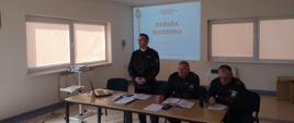 Zdjęcie przedstawia salę konferencyjną w której znajduje się stół prezydialny przy którym znajduje się trzech strażaków Państwowej Straży Pożarnej w umundurowaniu dowódczo-sztabowym. W tle slajdy prezentowane z rzutnika multimedialnego.