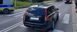 Czarny samochód osobowy, kombi, marki volvo, który stoi na jezdni, na pasach, na których doszło do potrącenia. Po lewej stronie na chodniku stoi radiowóz Policji.