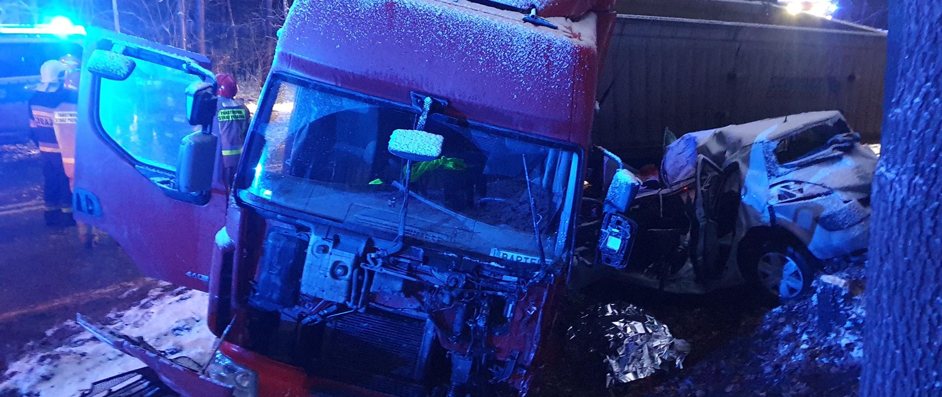 Samochód ciężarowy Renault Premium poza jezdnią , Renault Megan przygnieciony do drzewa przez ciężarowy, pora nocna, opady śniegu, w tle służby ratownicze
