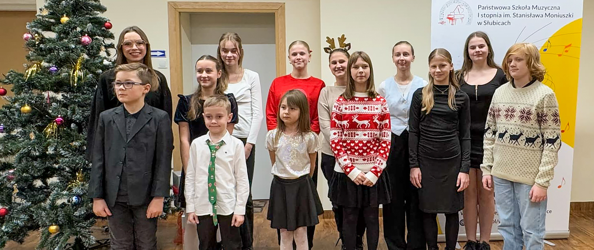 Na zdjęciu nauczycielka i kilkunastu uczniów stoją w auli szkolnej obok choinki.
