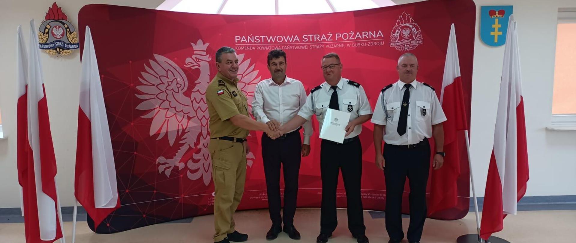 Komendant z 2 druhami w mundurach galowych na tle baneru straży pożarnej i flagami polski po bokach 