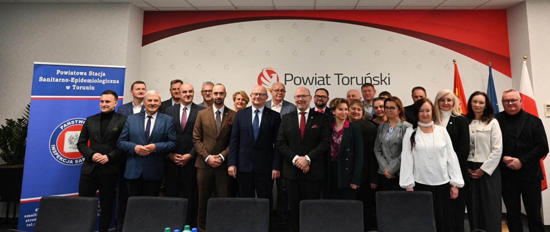 Grupa osób w strojach formalnych podczas oficjalnego wydarzenia w sali konferencyjnej Powiatu Toruńskiego, obok baneru PSSE i stojących flag