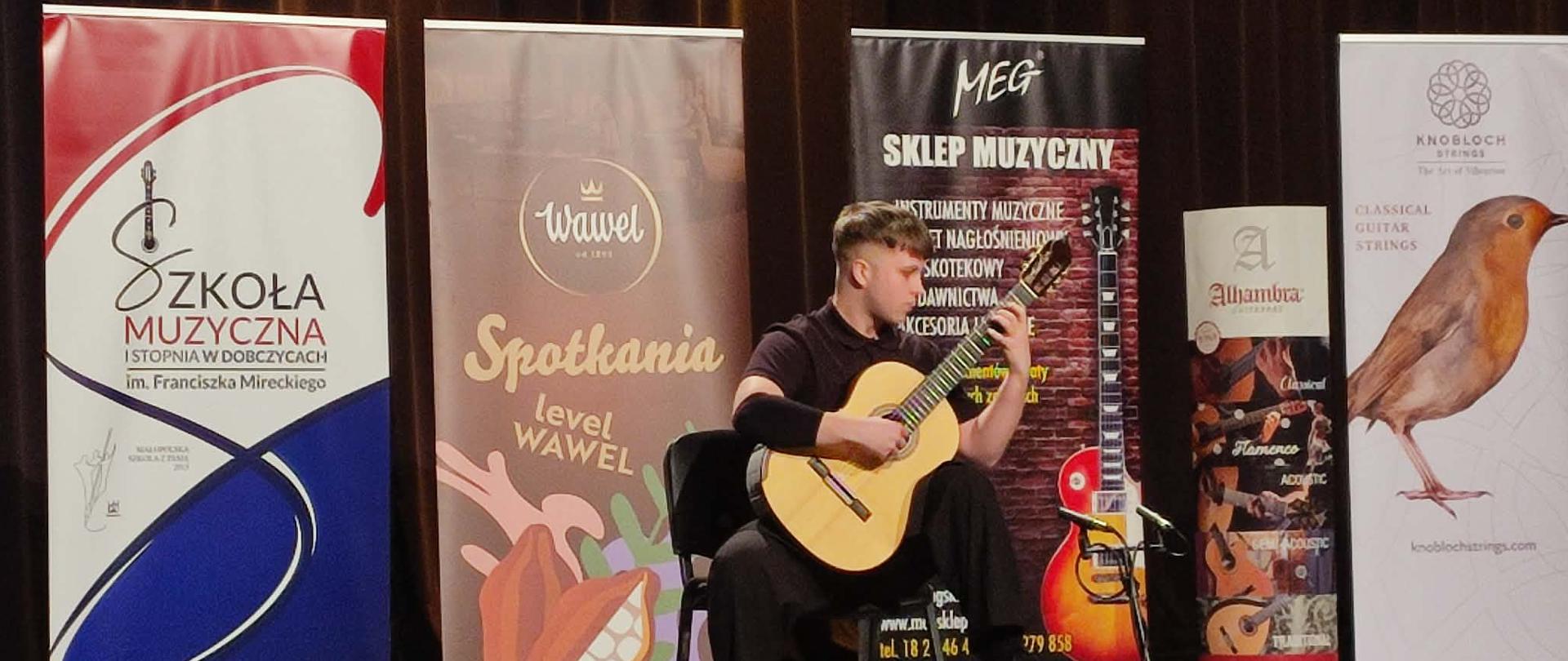 Na zdjęciu widzimy występ laureata I miejsca Bartłomieja Szypulskiego podczas VIII Ogólnopolskiego Konkursu Gitarowego „Młodzi Czarodzieje Gitary”, który odbył się 7 maja 2026 roku podczas koncertu finałowego w Szkole Muzycznej I stopnia w Dobczycach.
Młody gitarzysta prezentuje swój program artystyczny na scenie, wykonując utwór na gitarze klasycznej. Na scenie znajdują się również banery organizatorów i sponsorów konkursu.
Koncert laureatów był zwieńczeniem dwóch dni muzycznych zmagań młodych gitarzystów z całej Polski oraz okazją do zaprezentowania najlepszych wykonań nagrodzonych uczestników przed jury.
