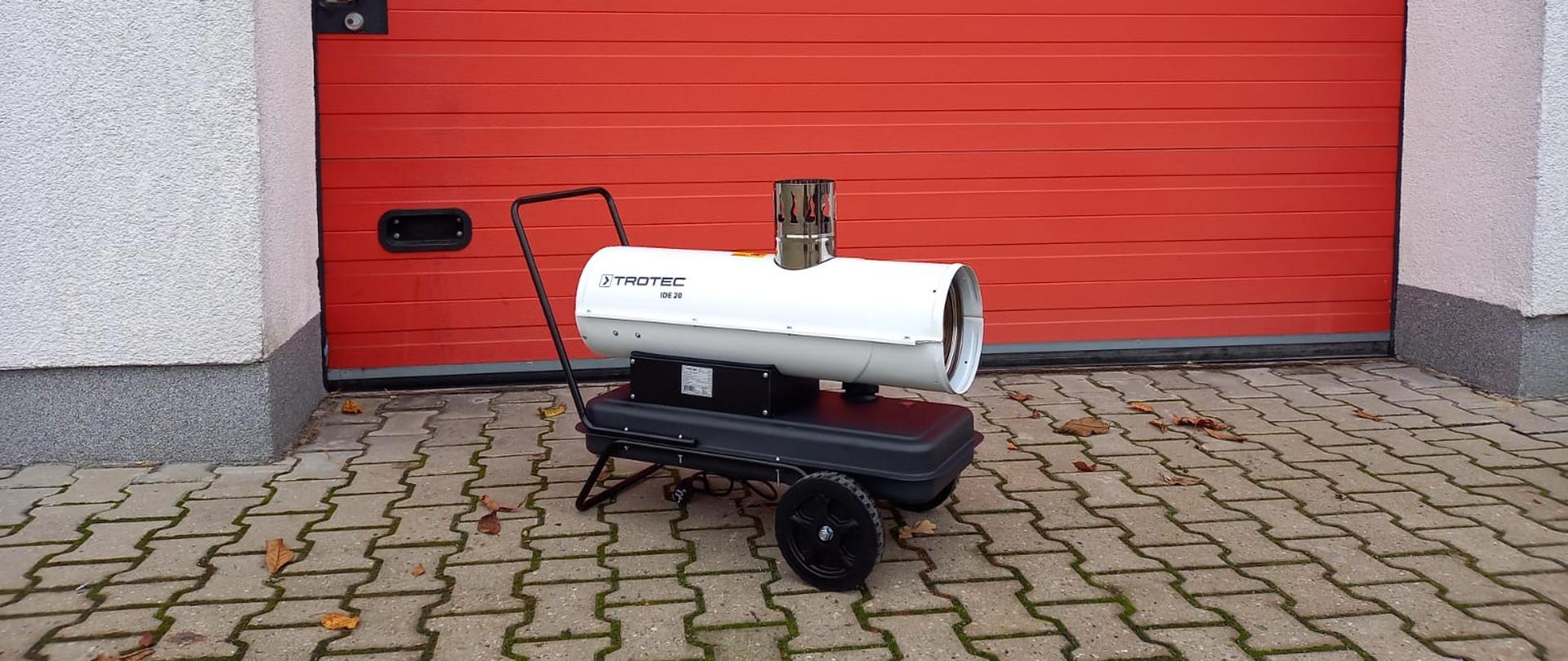 Zdjęcie przedstawia nagrzewnicę olejową koloru białego na tle czerwonej bramy garażowej Jednostki Ratowniczo-Gaśniczej Państwowej Straży Pożarnej w Strzelcach Krajeńskich