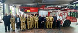 Grupowe zdjęcie uczestników wizyty studyjnej – polskich strażaków i ich niemieckich odpowiedników – wykonane w hali garażowej jednostki Feuerwehr Aachen, na tle nowoczesnego wozu strażackiego z drabiną mechaniczną.
