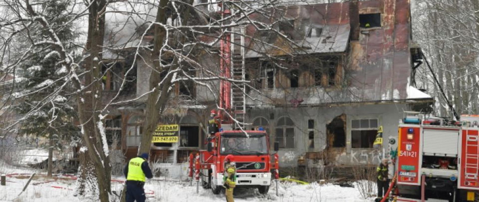 Spalony budynek piętrowy. Przed nim zastępy straty pożarnej porządkują teren. Wokół śnieg.