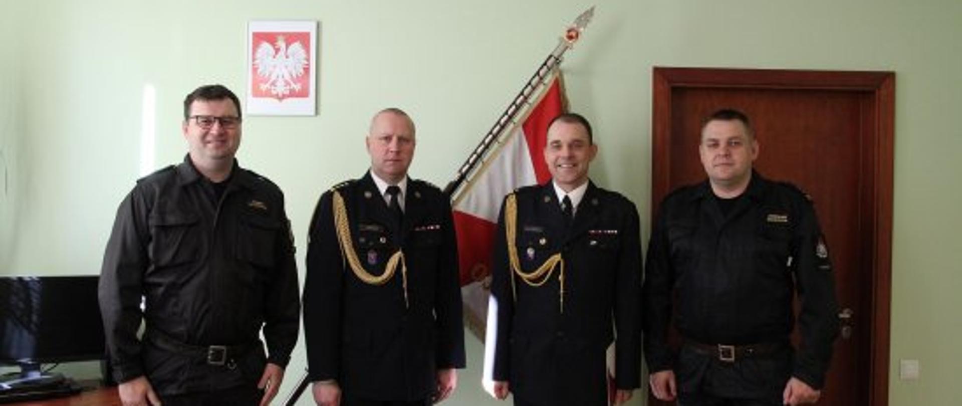 na tle zielonej ściany sztandaru oraz godła na ścianie stoi czterech strażaków na baczność, dwóch jest ubranych w ciemne mundury ze złotym sznurem, 