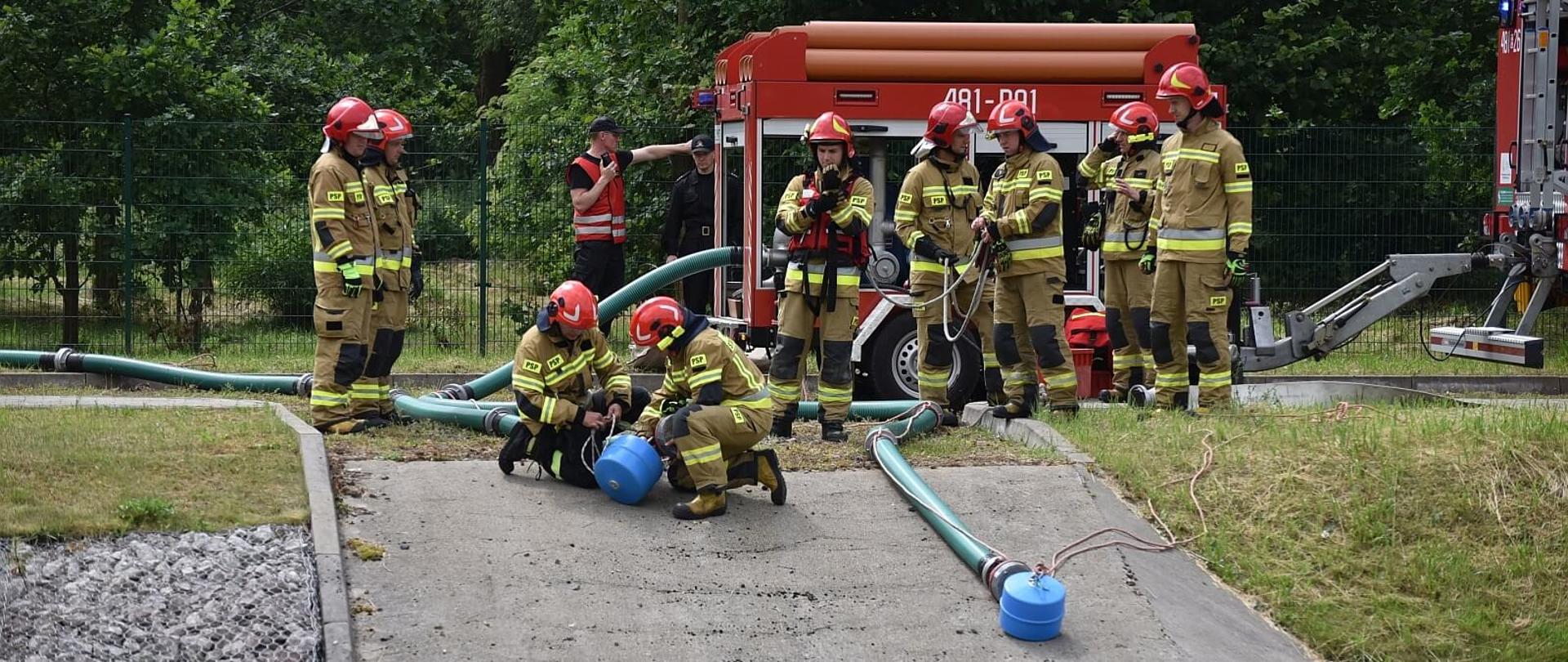 strażacy w ubraniach bojowych budujący linie ssawne za nimi przyczepa motopompą, w tle drzewa