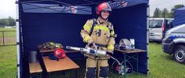 Nastoletni chłopiec stoi w specjalnym ubraniu strażackim i hełmie na tle namiotu Gminy Wadowice. W rękach trzyma ratownicze narzędzie hydrauliczne.