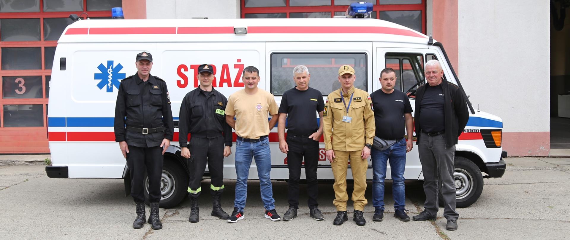 Grupowe zdjęcie Zastępcy Komendanta Centralnej Szkoły PSP w towarzystwie czterech druhów OSP oraz Naczelnika Wydziału Technicznego Centralnej Szkoły PSP i Zastępcy na tle przekazywanego samochodu specjalnego ambulansu