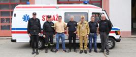 Grupowe zdjęcie Zastępcy Komendanta Centralnej Szkoły PSP w towarzystwie czterech druhów OSP oraz Naczelnika Wydziału Technicznego Centralnej Szkoły PSP i Zastępcy na tle przekazywanego samochodu specjalnego ambulansu