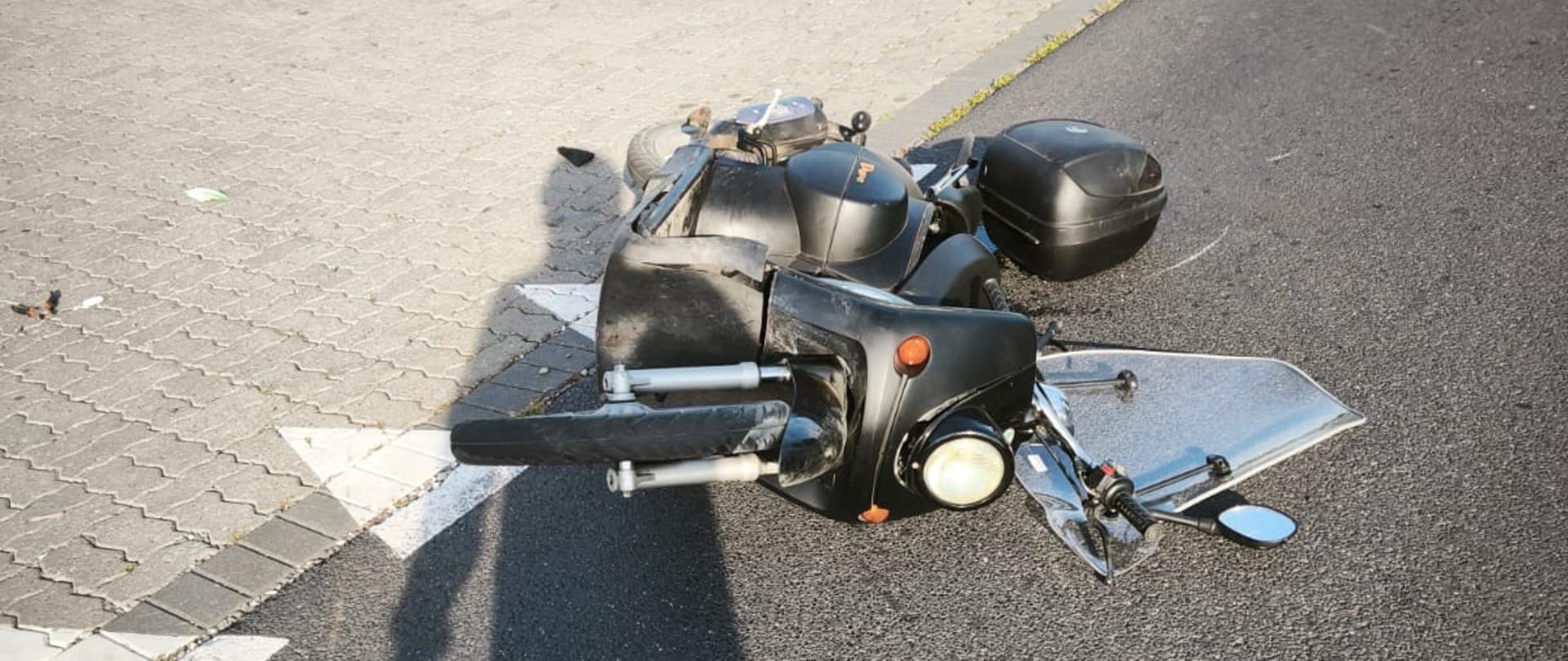 Na zdjęciu widać motocykl przewrócony na bok, leżący częściowo na asfaltowej jezdni i kostce brukowej przy poboczu. W tle znajduje się zielone pole z drzewami, a na pierwszym planie widoczny jest cień osoby robiącej zdjęcie.