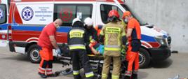 Na pierwszym planie ratownicy medyczni wraz ze strażakami PSP i OSP udzielają pomocy leżącej na noszach osobie poszkodowanej. W tle karetka ratowniczego pogotowia ratunkowego.