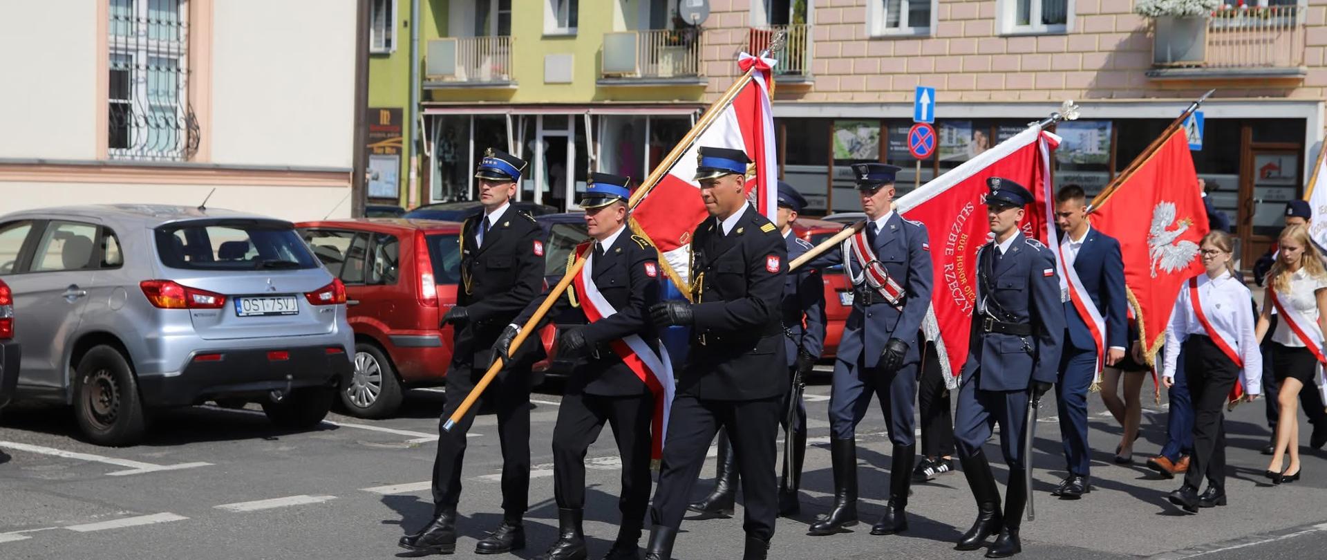Na zdjęciu znajdują się poczty sztandarowe, które idą w przemarszu, w tle widać budynki