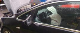 Samochód osobowy ciemnego koloru z podniesioną maską. Wewnątrz pojazdu w kierownicy zadziałała poduszka bezpieczeństwa.
