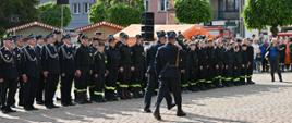 Strażacy stoją na płycie rynku w poododziale. Przed nimi przechodzi dwóch strażzaków i salutują. Za nimi w tle kamienice starego miasta. 