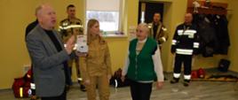 Wójt gminy omawiający zalety czujek czadu, obok seniorka, funkcjonariuszka PSP, w tle strażacy 