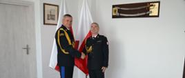 Pomorski komendant wojewódzki Państwowej Straży Pożarnej trzyma czerwoną teczkę oraz ściska dłoń strażaka za nimi stoją flagi Polski.