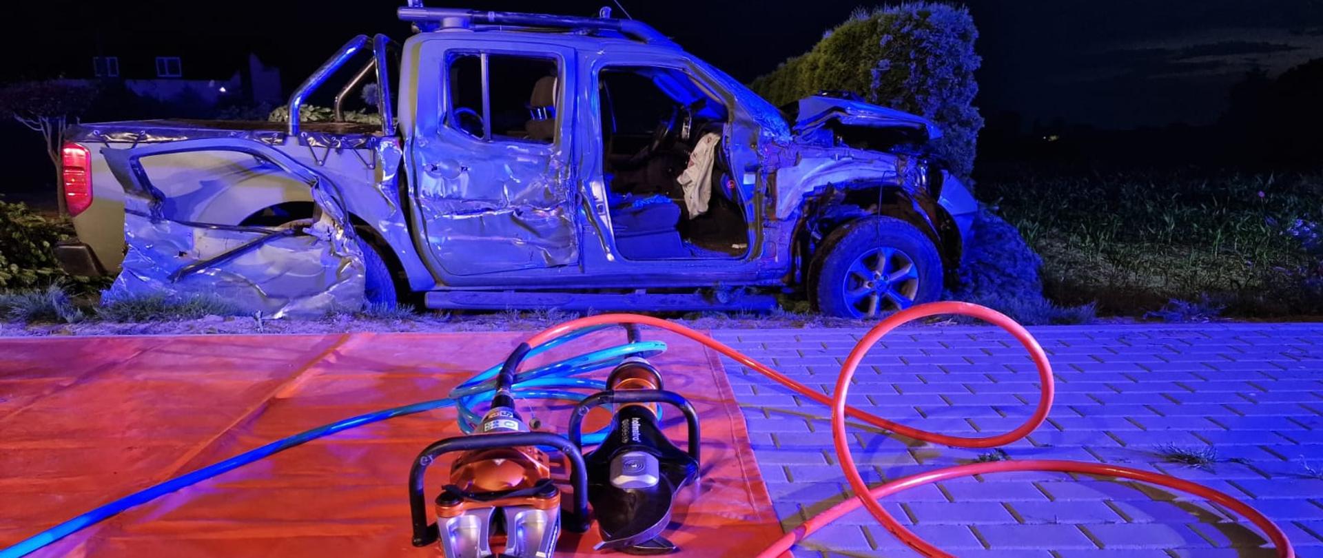 na zdjęci na pomarańczowej macie rozłożony sprzęt do ratownictwa drogowego w oddali samochód z pozbijany 