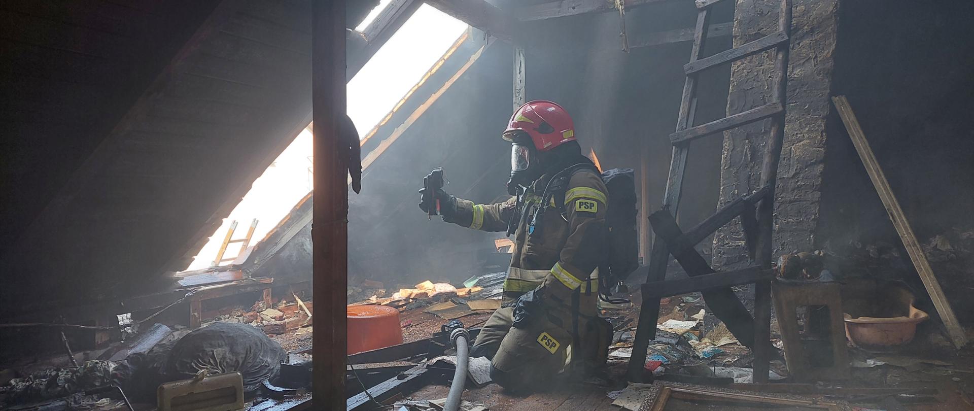 Zdjęcie przedstawia strażaka ubranego w ubranie specjalne koloru piaskowego oraz czerwony hełm. Na plecach nosi aparat ochrony dróg oddechowych, na twarzy natomiast ma założoną maskę oddechową od tego aparatu. Znajduje się w zadymionym strychu domu, gdzie był pożar. Strażak w dłoni trzyma kamerę termowizyjną. Obok znajduje się nawodniona linia gaśnicza zakończona prądownicą. Na podłodze znajdują się różne przedmioty. Po prawej stronie znajduje się komin, o który oparta jest stara drabina. W dachu budynku znajduje się duży otwór, przez który wpada światło dzienne.