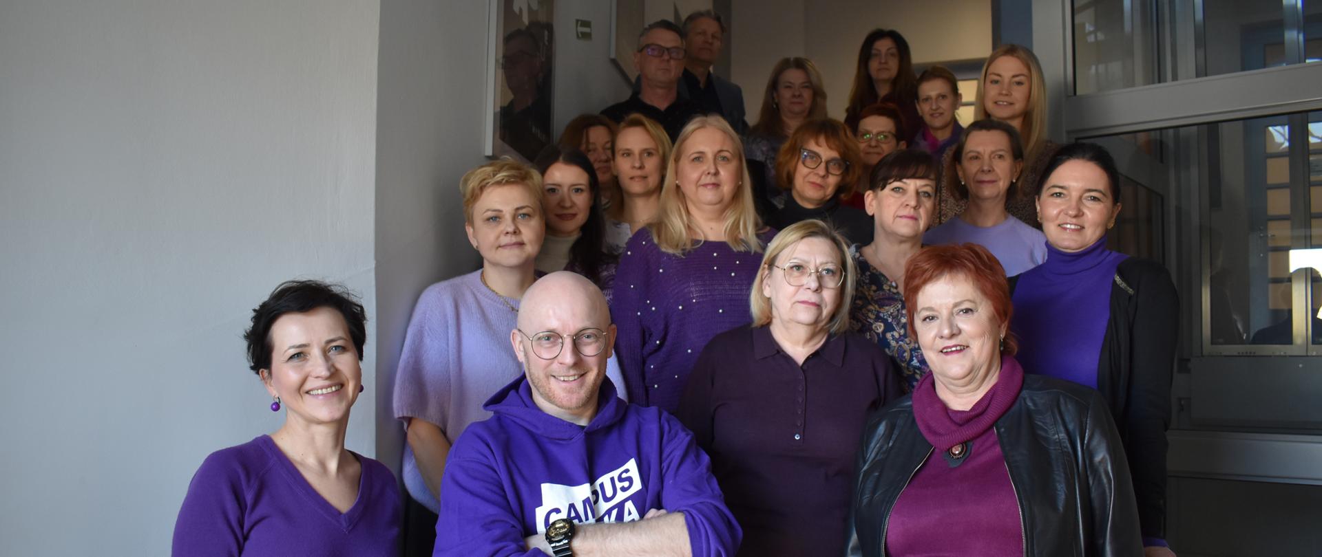 Widać większą grupę osób stojących na klatce schodowej wewnątrz budynku. Grupa ustawiona jest na kilku stopniach schodów, a część osób stoi również obok poręczy i przed wejściem do windy. Większość osób ma na sobie elementy garderoby w odcieniach fioletu, śliwki, purpury lub ciemnego bordo, ubranych z okazji Światowego Dnia Świadomości HPV. 