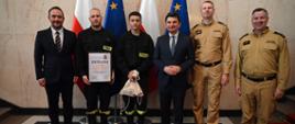 Sześć osób pozuje do pamiątkowego zdjęcia na tle flag Polski i Unii Europejskiej podczas wręczenia dyplomu i upominku dwóm strażakom.